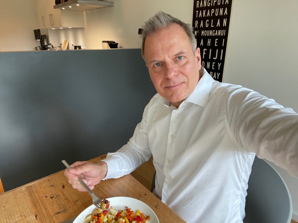 Ein Mann im weißen Hemd sitzt an einem Holztisch, genießt einen bunten Salat mit FRoSTA Tiefkühlprodukten und lächelt in seiner modernen Küche.