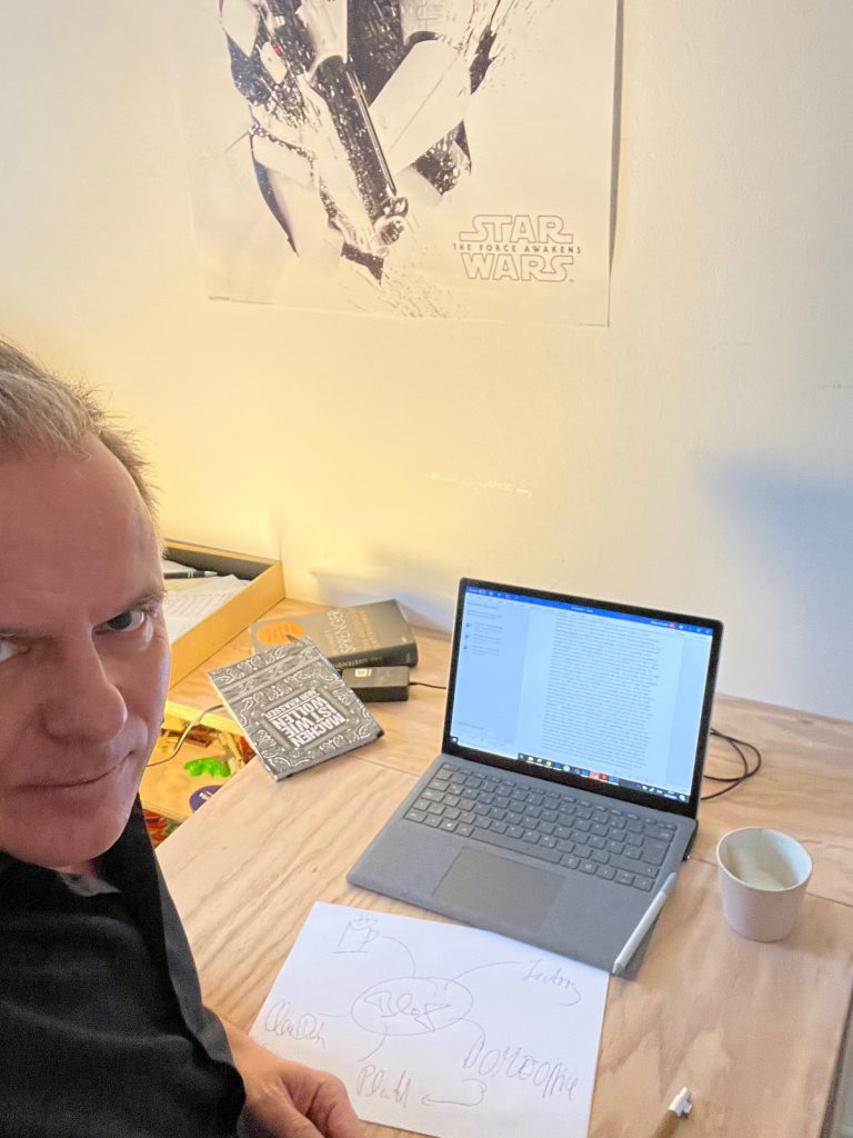 Eine Person sitzt an einem Schreibtisch mit Laptop, Büchern, einer Tasse, FRoSTA-Tiefkühlprodukten und Papieren. Ein Star Wars-Poster hängt hinter ihnen an der Wand.