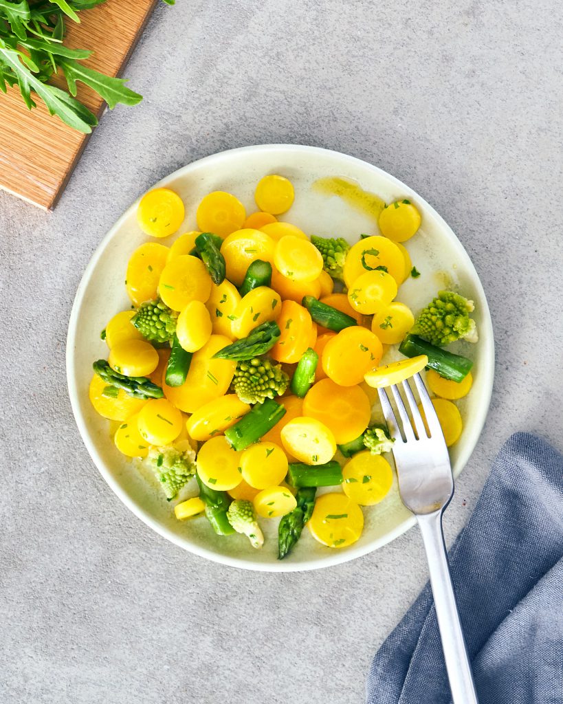 Ein Teller mit FRoSTA-Tiefkühlprodukten: gelbe Karotten, grüner Spargel und Romanesco, dazu Rucola, Gabel und graue Serviette.