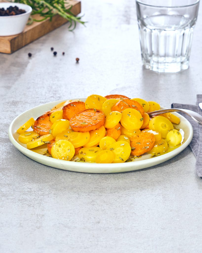 Ein Teller mit FRoSTA-Tiefkühlprodukten aus gelben und orangefarbenen Karotten, verfeinert mit Kräutern, liegt neben Besteck und Wasser.