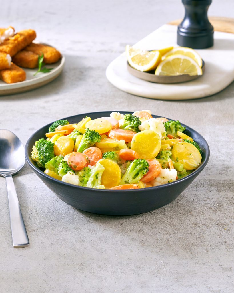 Eine schwarze Schüssel mit FRoSTA Tiefkühl-Königsgemüse wie Karotten, Brokkoli und Kartoffeln steht neben FRoSTA Fischstäbchen und Zitronenspalten.