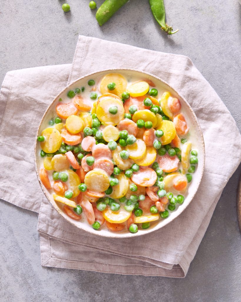 Eine Schüssel cremiges FRoSTA-Mischgemüse – Karotten, Kartoffeln, grüne Erbsen – ist die perfekte FRoSTA-Gemüsebeilage auf beigem Tuch.