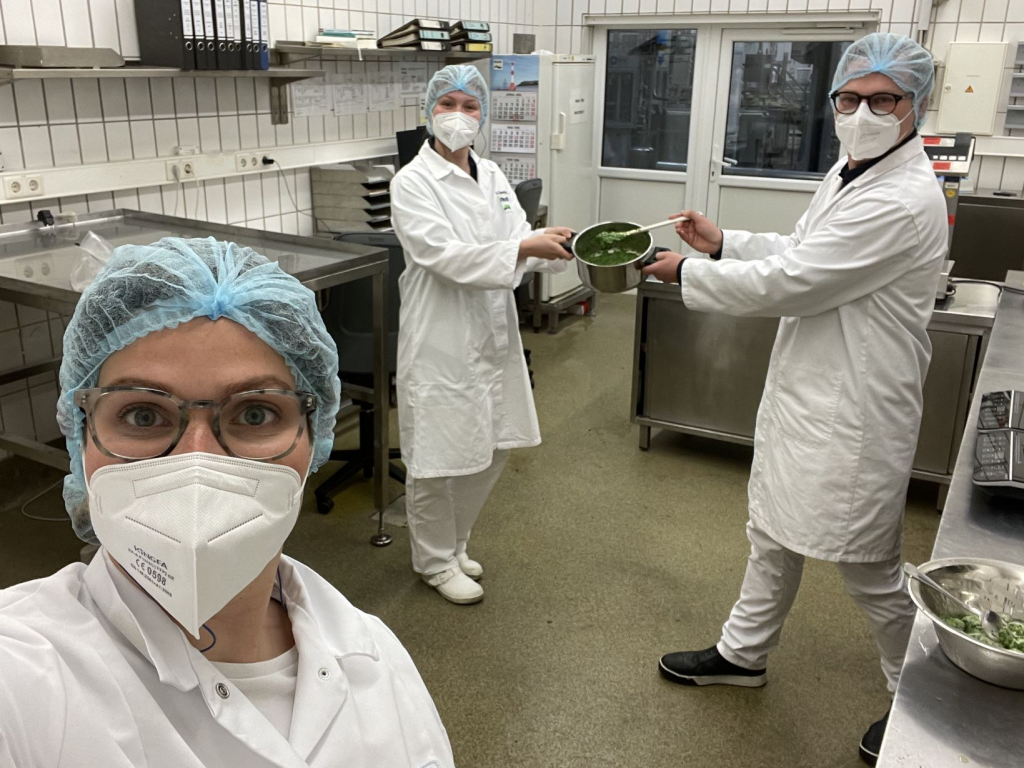 Drei Personen in Laborkitteln machen ein Selfie in einer sauberen Küche der Spinaternte Bobenheim-Roxheim, halten eine Schale FRoSTA-Tiefkühlprodukte.