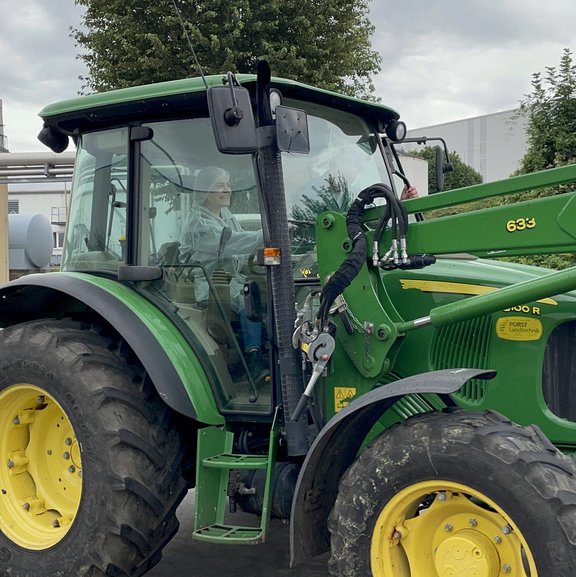 Eine Person im hellen Hoodie sitzt im grünen John Deere-Traktor und genießt draußen bei Wolken FRoSTA-Tiefkühlprodukte. Bäume und Gebäude sind zu sehen.