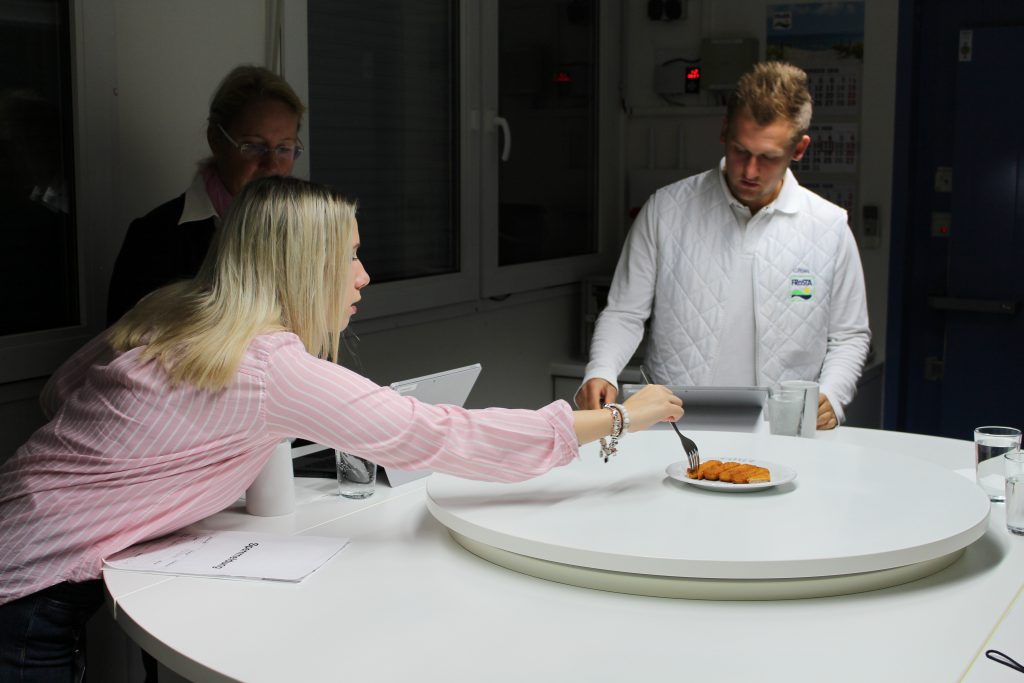 Drei Personen stehen bei einer Ausbildung um einen Tisch. Eine Frau greift nach FRoSTA-Tiefkühlprodukten, während die anderen zuschauen.