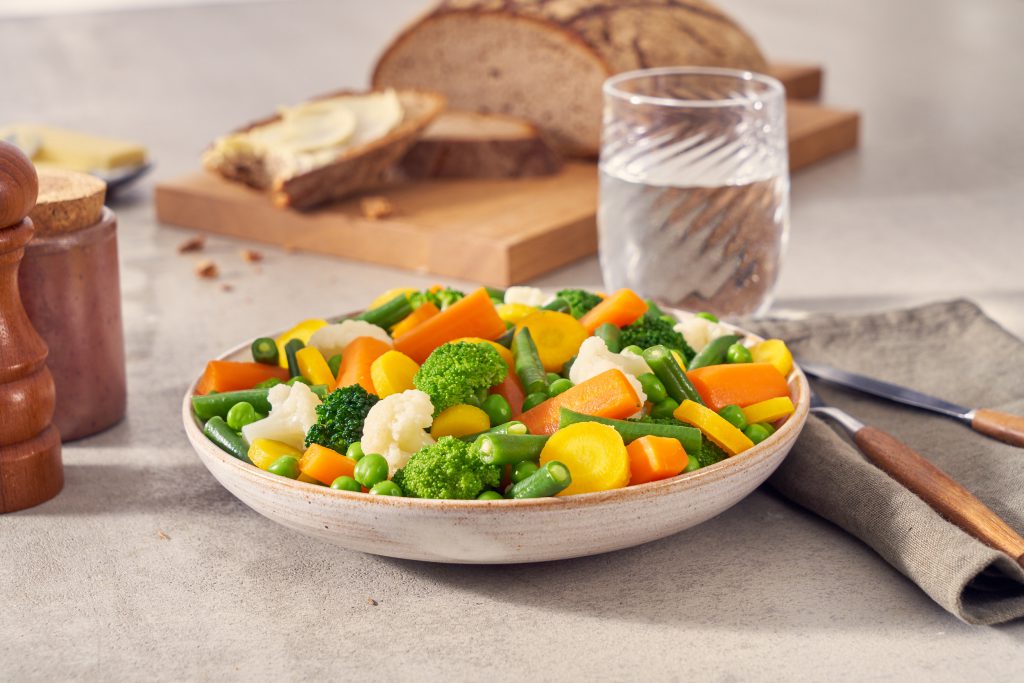 Eine Schüssel FRoSTA Gemüse Mix mit gedünsteten Karotten, Brokkoli, Blumenkohl, Erbsen und grünen Bohnen steht mit Brot und Wasser auf einem Tisch.